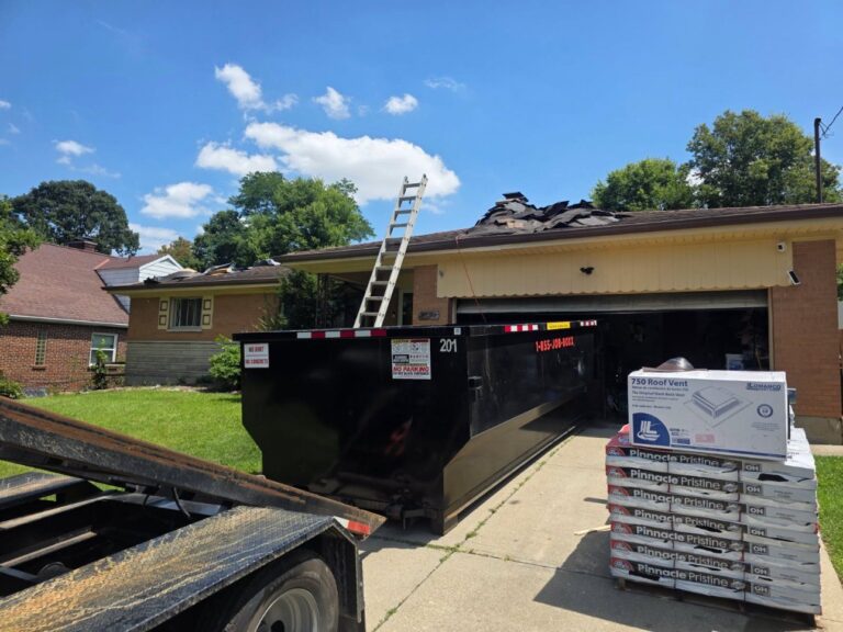 Roll-off dumpster on a driveway during a roof tear-off, with a ladder up to the stripped roof