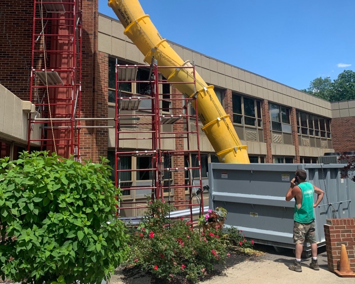 Yellow sectional trash chute hanging from red scaffolding down a two-story brick building into a grey roll-off dumpster, with a worker standing beside it